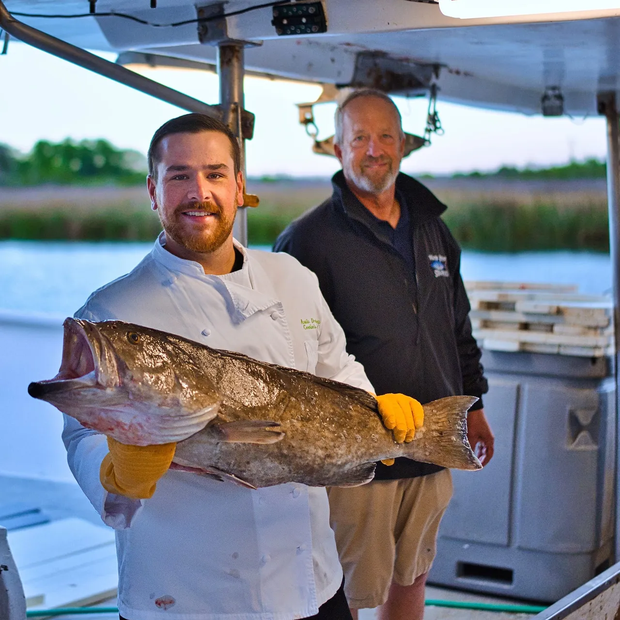 A visiting chef holding a large grouper beside a member of the Water Street team aboard a boat